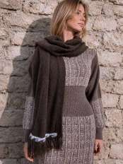 Blonde woman by a stone wall wearing a knitted dress and a brown fringed alpaca wool shawl wrap, model Kora.